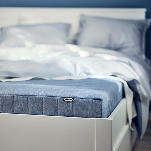 Primer plano del colchón VADSÖ azul-gris de ikeas sobre cama blanca, mostrando la funda de tela y la tapa de espuma.