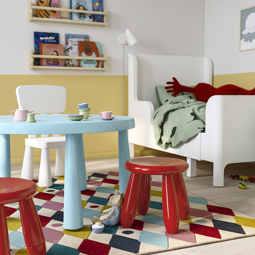 Zona de juegos infantiles con mesa azul, taburetes rojos. Alfombra brillante, silla blanca.