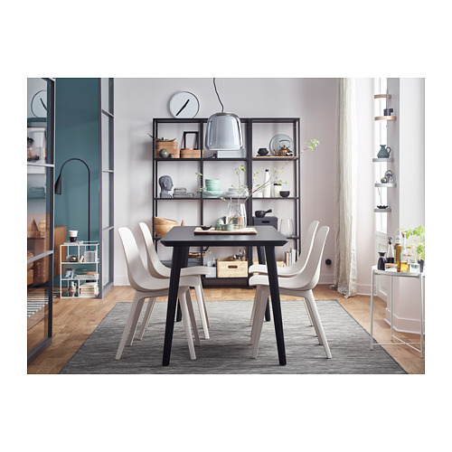 Moderno espacio de comedor con mesa negra, cuatro sillas blancas, alfombra gris y estantes de vidrio con marco negro que muestran la decoración.