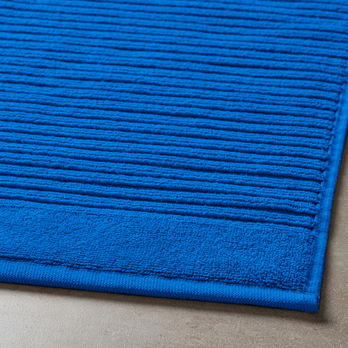 Alfombrilla de baño azul brillante, hecha de algodón suave y absorbente, esquina desplegada sobre un piso gris, mostrando su superficie texturizada.