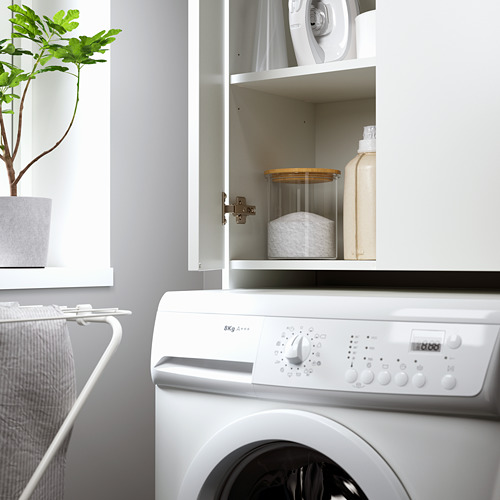 Moderna instalación de lavandería con lavadora, estante abierto con detergente y paños, y planta en maceta, ideal para espacios pequeños.