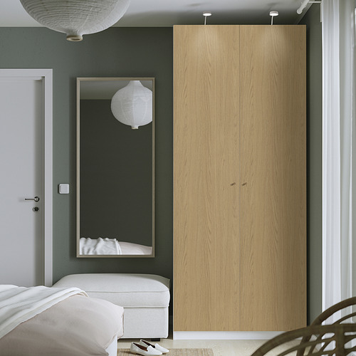 Un dormitorio moderno, un armario alto de madera clara PAX junto a un gran espejo y una cama blanca.