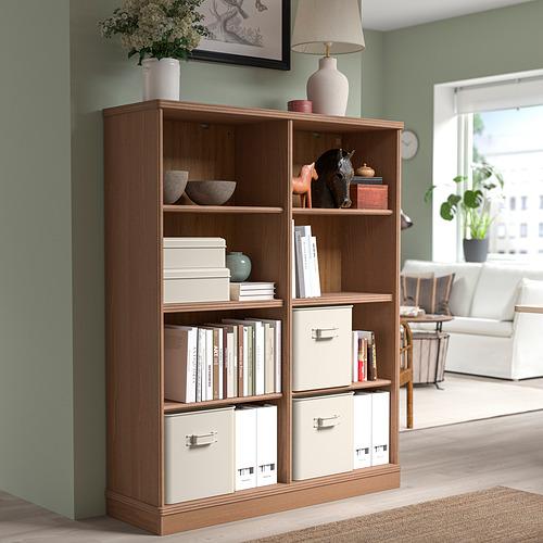 Librería LANESUND de color marrón claro con libros, decoración y almacenamiento. Lámpara alta y gran cuadro encima. Sala luminosa y abierta con comedor y sofá.