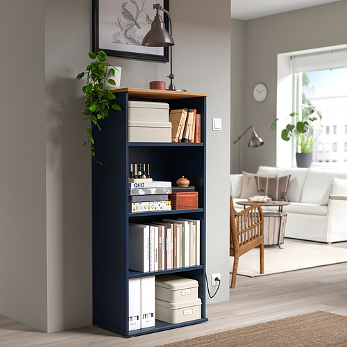 Estantería PLOGFÅRA azul oscuro con libros, cajas, plantas y lámparas en un acogedor espacio habitable.