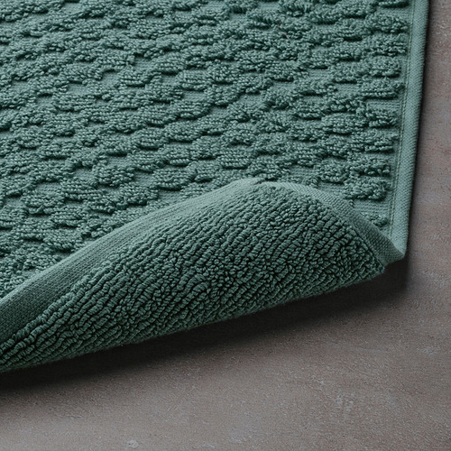 Alfombra de baño verde texturizada en primer plano, mostrando su rizo suave y su alta absorción. La esquina ajustada revela un diseño lujoso.