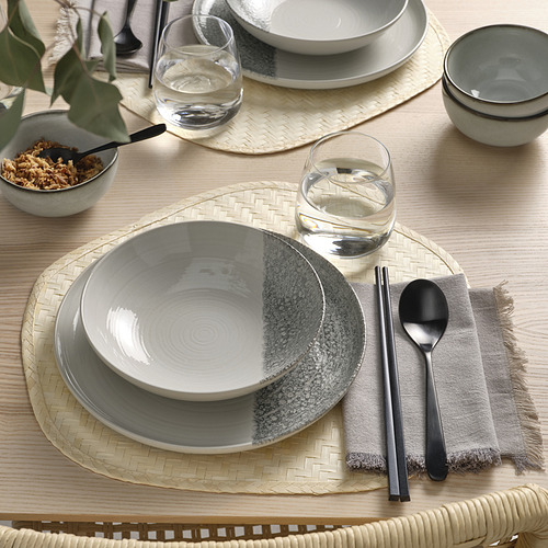 Juego de mesa con platos KRUSTISTEL, con diseños grises y blancos. Incluye tazones, vasos y cubiertos dispuestos en manteles individuales tejidos.