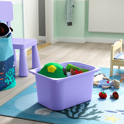 Contenedor de almacenamiento de juguetes de lavanda que contiene bloques de colores y una serpiente de juguete sobre una alfombra azul con una silla y una bolsa de juguete en un área de juegos para niños.