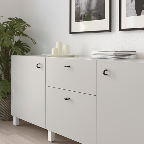 Mueble blanco con dos puertas y cajones, con tiradores y patas de metal negro. Decorado con velas y revistas apiladas en la parte superior.