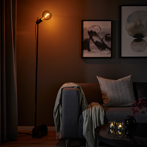 Acogedor rincón con lámpara de pie negra HÅRSLINGA, sofá gris, cojines, mantas y velas encendidas sobre la mesa con arte en la pared.