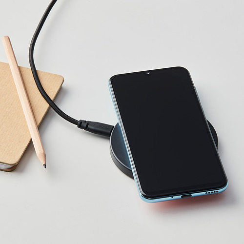 Almohadilla de carga inalámbrica LIVBOJ negra con teléfono inteligente, cable y computadora portátil sobre una superficie ligera. Diseño compacto y delgado.