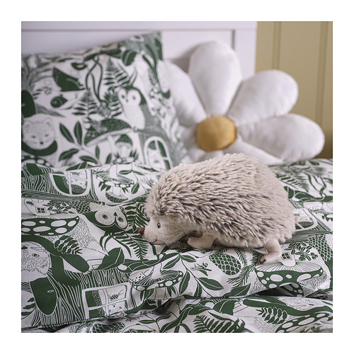 Un erizo de peluche SKOGSDUVA gris sobre un lecho de flores verdes. Es pequeño, redondo y afelpado, colocado entre almohadas grandes.