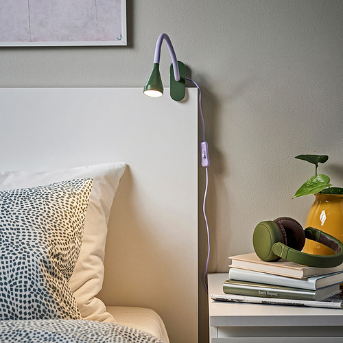 Acogedor dormitorio con lámpara de color verde púrpura, mesita de noche, libros, auriculares, planta.