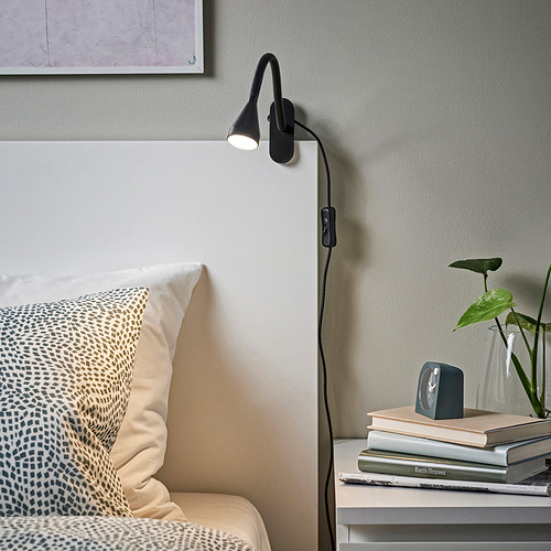Lámpara NÄVLINGE negra sobre cabecero blanco, mesita de noche iluminada con libros y plantas. Almohada estampada en la cama.