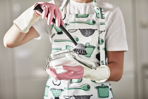 Una persona con un delantal blanco usa un cepillo de fregar RINNIG verde y blanco para limpiar un frasco de vidrio en la cocina.