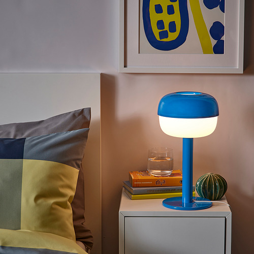 Mesita de noche con lámpara SOLHETTA azul, libros y cristal. Almohadas de colores y arte en la pared.