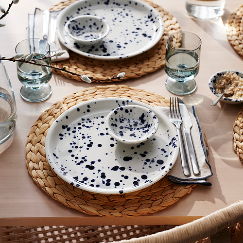 Mesa con platos, cubiertos, manteles individuales tejidos y vasos moteados de azul, listos para comer.
