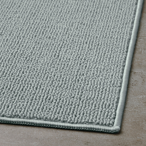 Alfombrilla de baño gris claro con una superficie de tela texturizada y en bucle y un borde liso, que se muestra sobre una superficie plana.