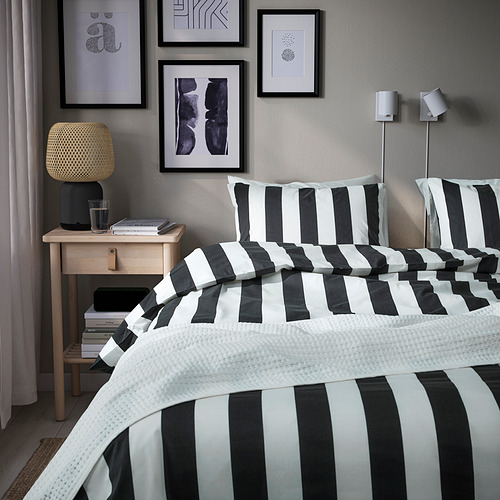 Dormitorio moderno con cama a rayas blancas y negras, lámpara amarilla, libros, arte, almohada de calabaza, luz acogedora.