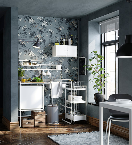 Cocina moderna con papel tapiz floral, nevera pequeña y carritos de almacenamiento. Decoración blanca y acentos negros, gran ventanal, que añade luminosidad.