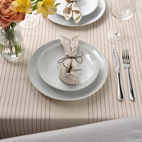 Mesa con platos blancos, servilletas dobladas, cubiertos, copa de vino y mantel a rayas; flores en jarrón.