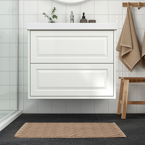 Baño moderno con mueble blanco, alfombrilla FJÄLLKATTFOT beige y taburete de madera. La alfombrilla es lujosa, cabe debajo del gabinete y agrega calidez.
