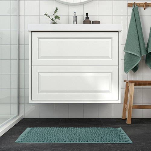 Un baño con un gabinete y lavabo blancos, alfombra de baño verde, toallas y plantas.