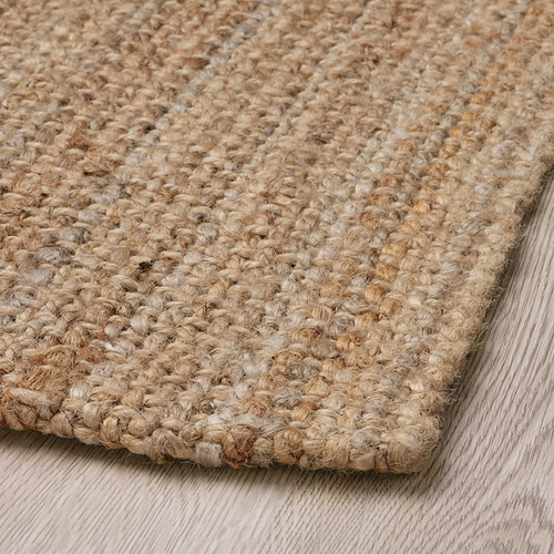 Alfombra de yute natural en primer plano, mostrada sobre suelo de madera. Tejido denso, superficie texturizada, robusto pero elegante.