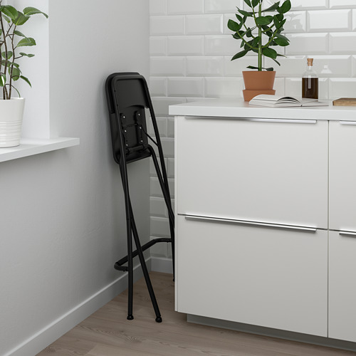 Taburete plegable negro con reposapiés, apoyado en armarios blancos en una cocina. Fácil de almacenar y usar para asientos adicionales.