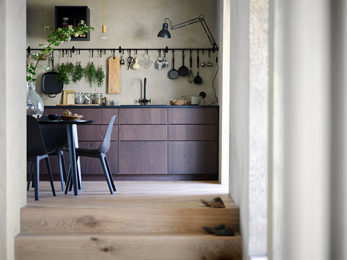 Cocina moderna con armarios de madera oscura, ollas colgantes y una mesa de comedor negra con sillas. Una lámpara y plantas decoran el espacio.