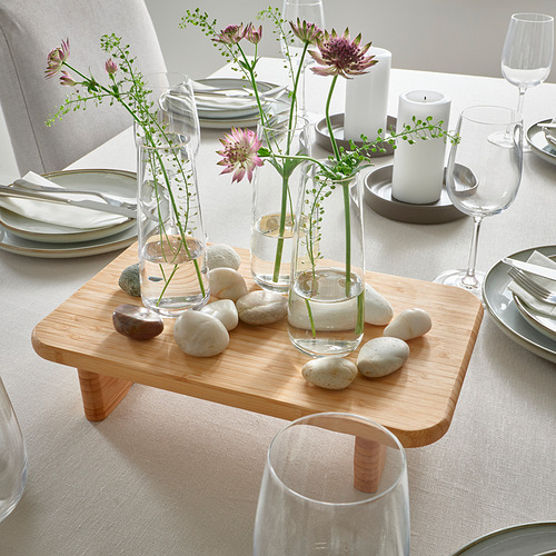 Plataforma elevada de madera que muestra tres jarrones de vidrio con flores moradas y guijarros, rodeados de platos, vasos y velas sobre una mesa.