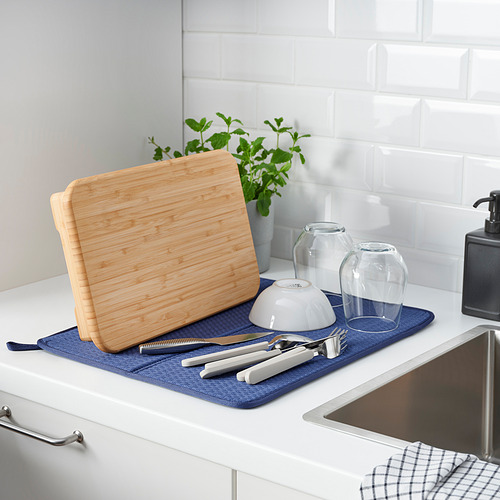 Escena de cocina con tabla de cortar, cuchillos, tazón, jarrones y hierbas en una estera azul cerca del fregadero.