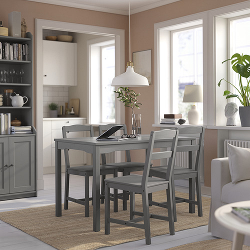 Comedor luminoso con mesa y sillas de madera gris, lámpara colgante blanca, alfombra de yute y estantería con accesorios, junto a una cocina moderna.
