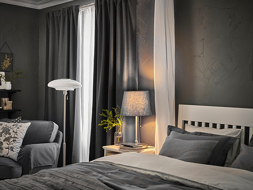 Dormitorio con paredes negras, cama blanca con almohadas grises, cortinas grises, lámpara gris con pantalla blanca SKOTTORP, y sillón gris junto a la ventana.