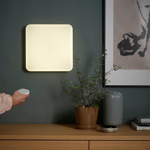 Una mano apunta con un control remoto a un panel de luz cuadrado montado en la pared, controlando su brillo y color, junto a una planta de interior y obras de arte.