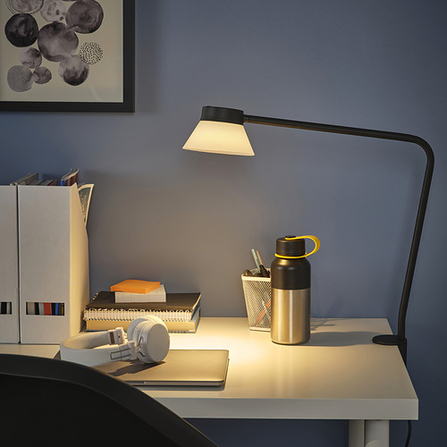 Lámpara SAMBORD de sobremesa con pantalla triangular, que ilumina el espacio de trabajo con libros, botellas y auriculares.