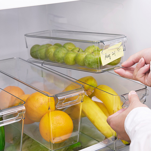 Una mano que accede a los cajones transparentes de la nevera con frutas. El cajón superior tiene una nota: 21 de agosto.