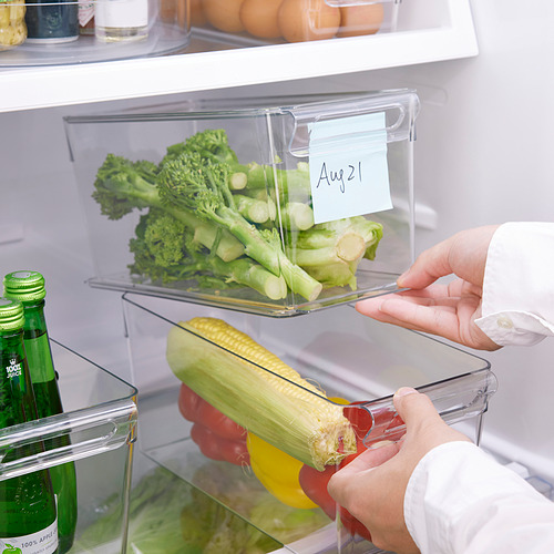 Una persona organiza los productos en un refrigerador usando cajas de almacenamiento transparentes y apilables. Una nota con fecha se fija debajo del mango.
