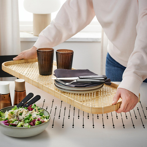 Persona que lleva una bandeja de bambú con platos, cubiertos y vasos, y coloca la ensaladera sobre la mesa.