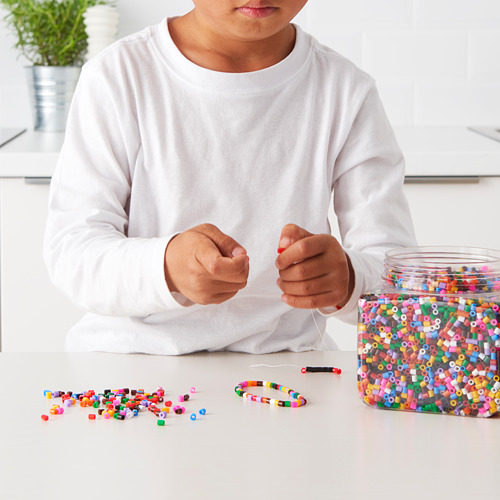 Niño ensartando cuentas de colores en una mesa blanca. Los tarros de cuentas y patrones muestran la motricidad fina y la creatividad.