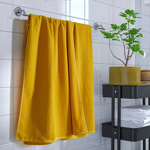 La toalla GULVIAL de color amarillo brillante cuelga cerca de una pequeña unidad de almacenamiento negra con una planta verde y toallas dobladas, que contrastan con los azulejos blancos.