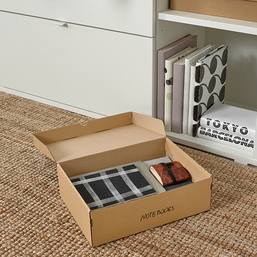Caja de cartón con libros de notas etiquetados en el suelo junto a un estante blanco, con libros de notas y una billetera marrón.