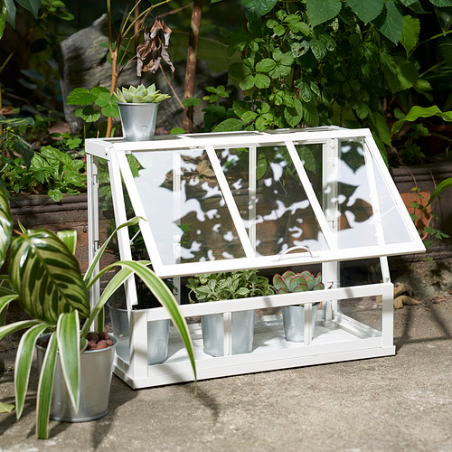Un mini invernadero blanco con macetas metálicas que albergan las plantas tanto en el interior como en la parte superior, proporcionando protección y fácil acceso para el crecimiento.