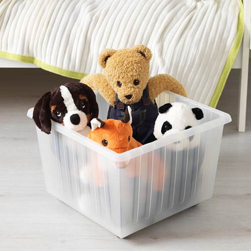 Un contenedor de almacenamiento de plástico transparente con ruedas, que sostiene varios animales de peluche, se encuentra en un piso de madera junto a una cama.
