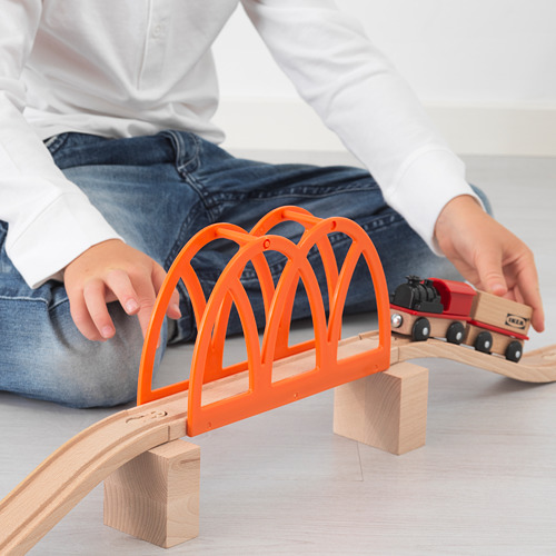 Niño sentado en el suelo, jugando con un tren de madera. El puente de color naranja brillante cruza las vías, el tren lo cruza, promoviendo la imaginación y las habilidades motoras.