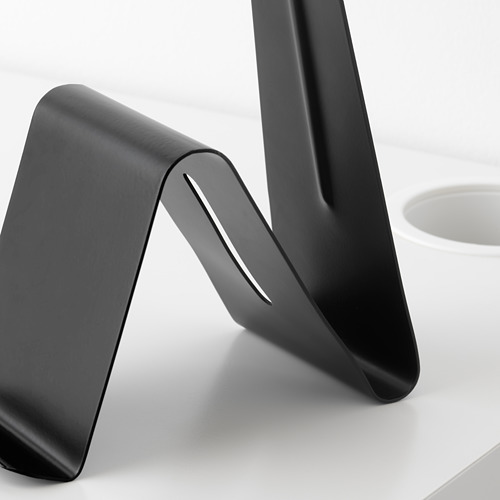 Soporte de metal negro con bordes lisos y redondeados; sostiene tabletas o teléfonos, se sienta en un escritorio blanco.