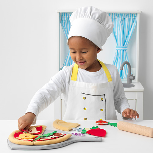 Niño jugando con un juego de pizza de juguete, con gorro y delantal de chef, colocando aderezos de fieltro en la base de la pizza.
