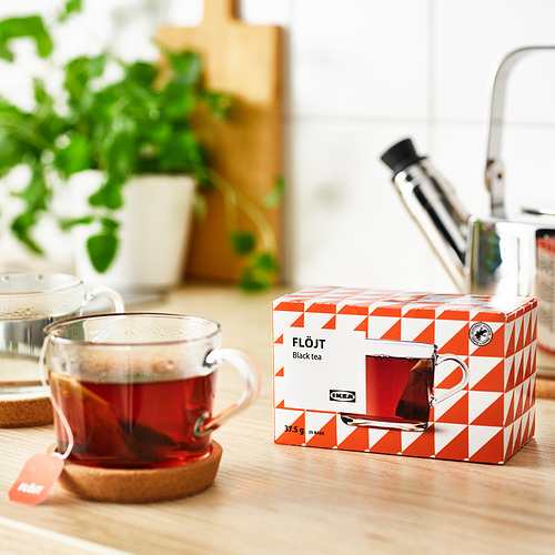 Taza transparente de té negro humeante junto a una caja de té FLÖJT de ikea abierta con 25 bolsitas de té sobre una mesa de madera, lo que sugiere que está lista para beber.