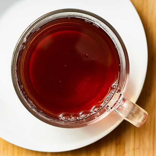 Taza de vidrio transparente llena de té rojo oscuro en un platillo blanco. Bebida calmante y humeante con rico sabor.