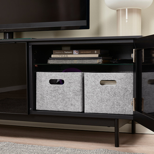 Mueble de TV negro con puertas abiertas, cajas de almacenamiento de fieltro gris, libros y una lámpara.
