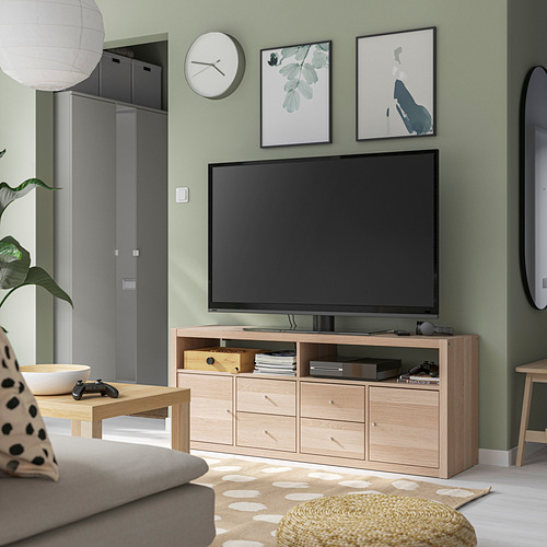 Salón moderno con mueble de tv de roble claro KALLAX. Televisor de pantalla plana, controladores de juegos, paredes verdes, almohadas y alfombra estampada.